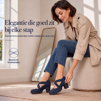 Smiling woman seated on a pouffe adjusting the strap of a navy suede-look block heel, with a mirror behind. 4