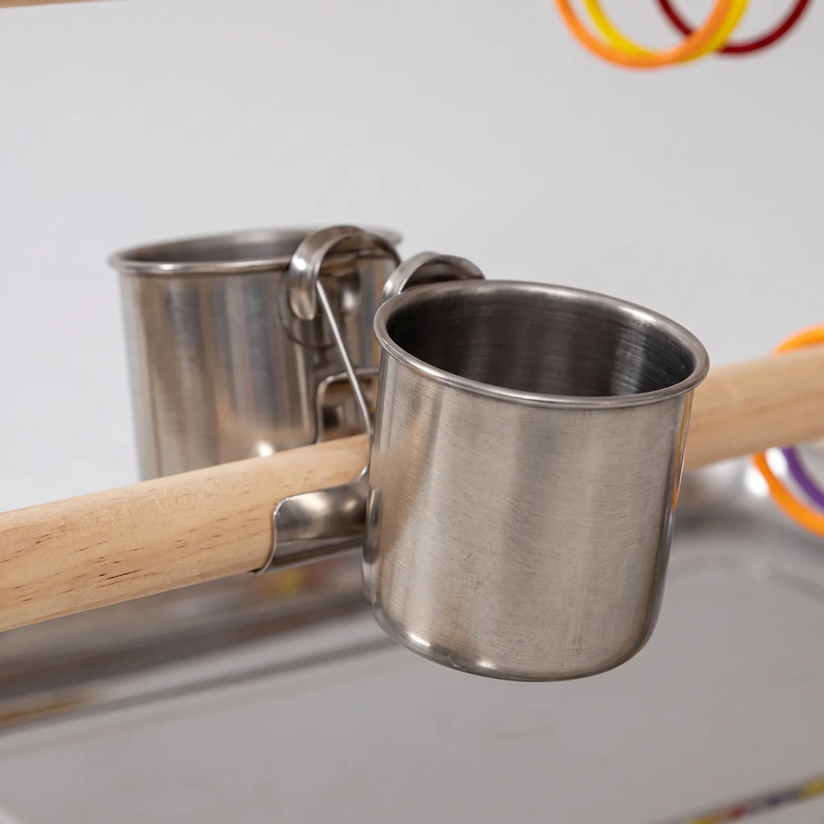 Close-up of two stainless steel feeding cups clipped onto a wooden perch above the metal tray, with coloured rings behind. 3
