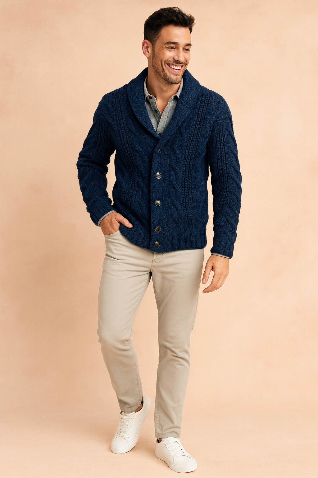 Full-length view of a man wearing the navy cardigan over a grey shirt with beige trousers and white trainers on a peach background. 2