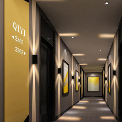 Black wall lights spaced along a hotel corridor, casting narrow vertical beams of warm light on grey walls 2