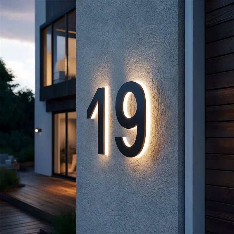 Black number 19 with a warm halo of backlight on a grey rendered wall at dusk, beside a timber-clad house. 3