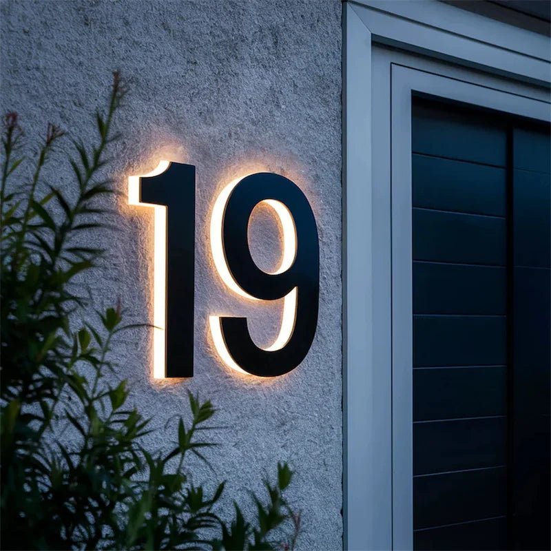 Close view of a black number 19 with glowing backlight on a rough grey wall beside a dark door and plant. 5
