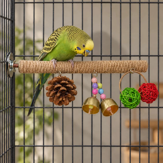Green and yellow budgerigar standing on the rope-wrapped perch inside a cage, looking down at the pine cone, bells and woven balls. 2
