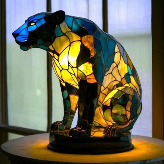 Seated panther lamp in blue, green and yellow glass on an oval base, placed in front of a window. 16