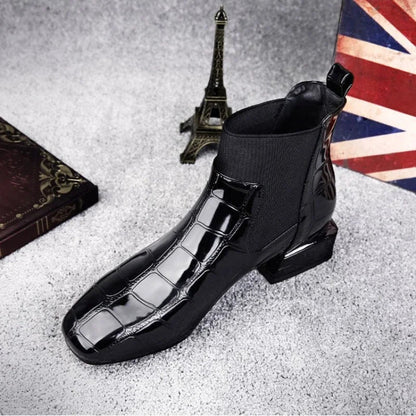 Black glossy croc-effect ankle boot with square toe and elastic side panel, seen from above-front on grey carpet beside a model Eiffel Tower. 1