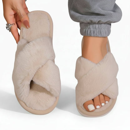 Cream fluffy crossover slippers, one worn with grey cuffed joggers and the other held up showing the plush footbed. 7