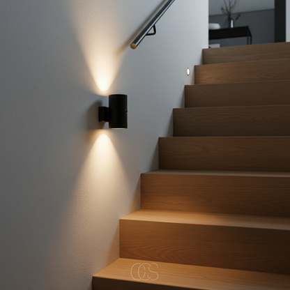 Black cylindrical light on a pale wall beside wooden stairs and a handrail, throwing warm beams upward and downward. 5