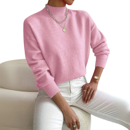 Model seated on a cream armchair wearing the jumper in pink with white jeans, layered silver necklaces and a gold watch. 1