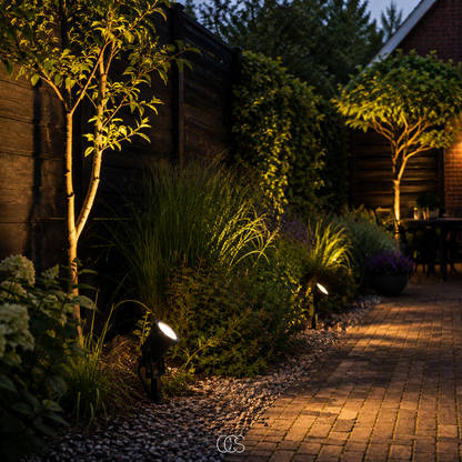 Several spotlights along a gravel border at night, lighting grasses, shrubs and trees beside a brick-paved garden path. 4