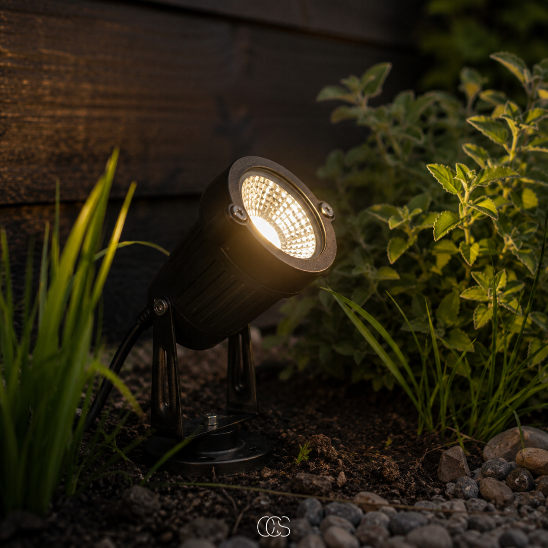 Black spotlight lit with a warm glow at dusk, set in dark soil beside grass, leafy herbs and pebbles. 1