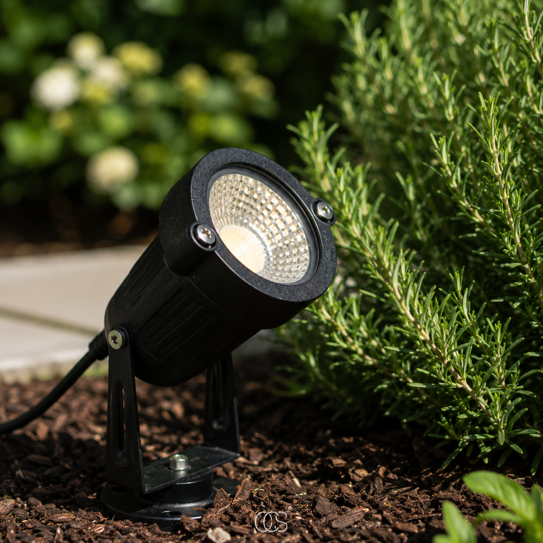 Close-up in daylight of the black spotlight on its base in bark mulch, beside a rosemary bush and a paving edge. 2