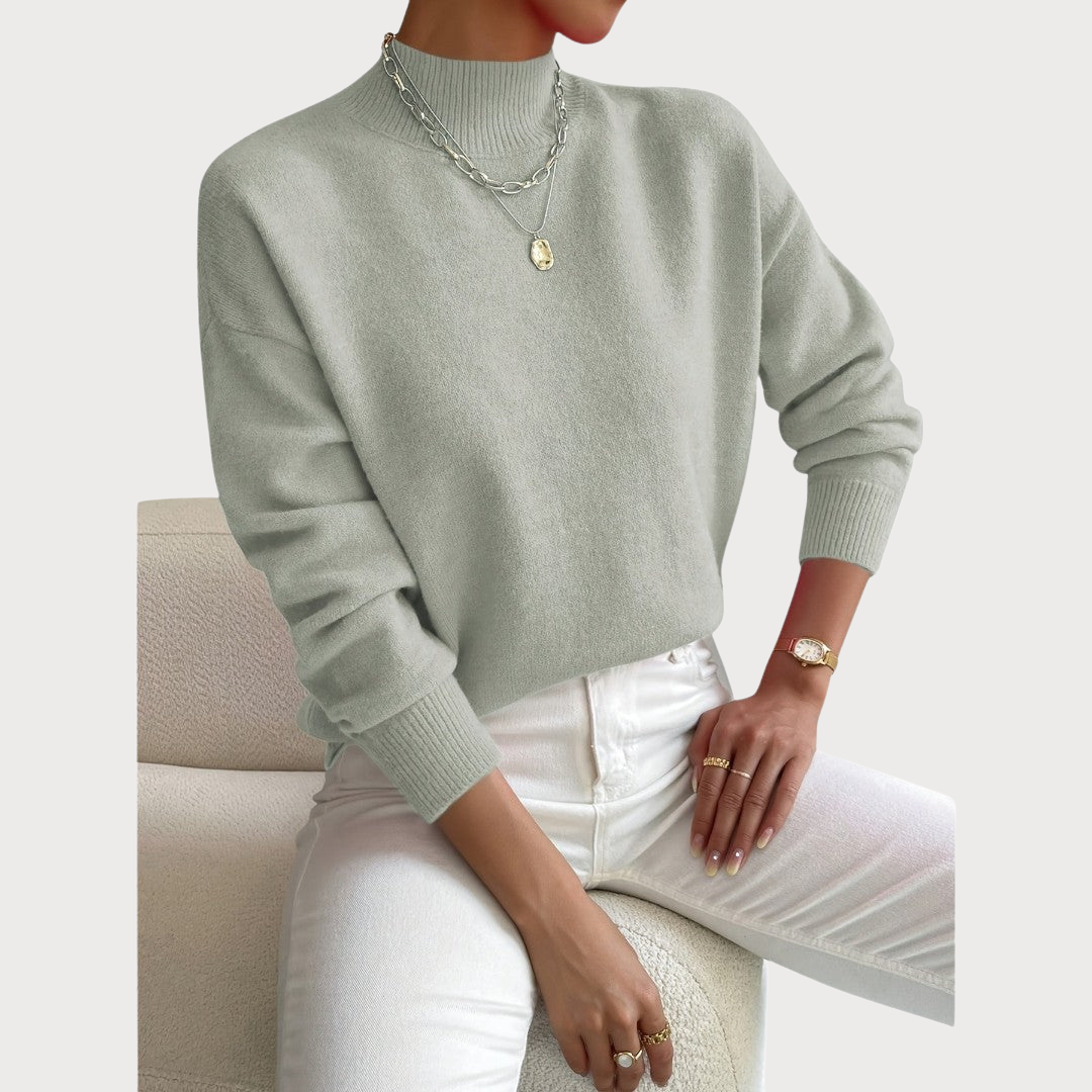 Model seated on a cream armchair wearing the jumper in soft sage green with white jeans and layered necklaces. 2
