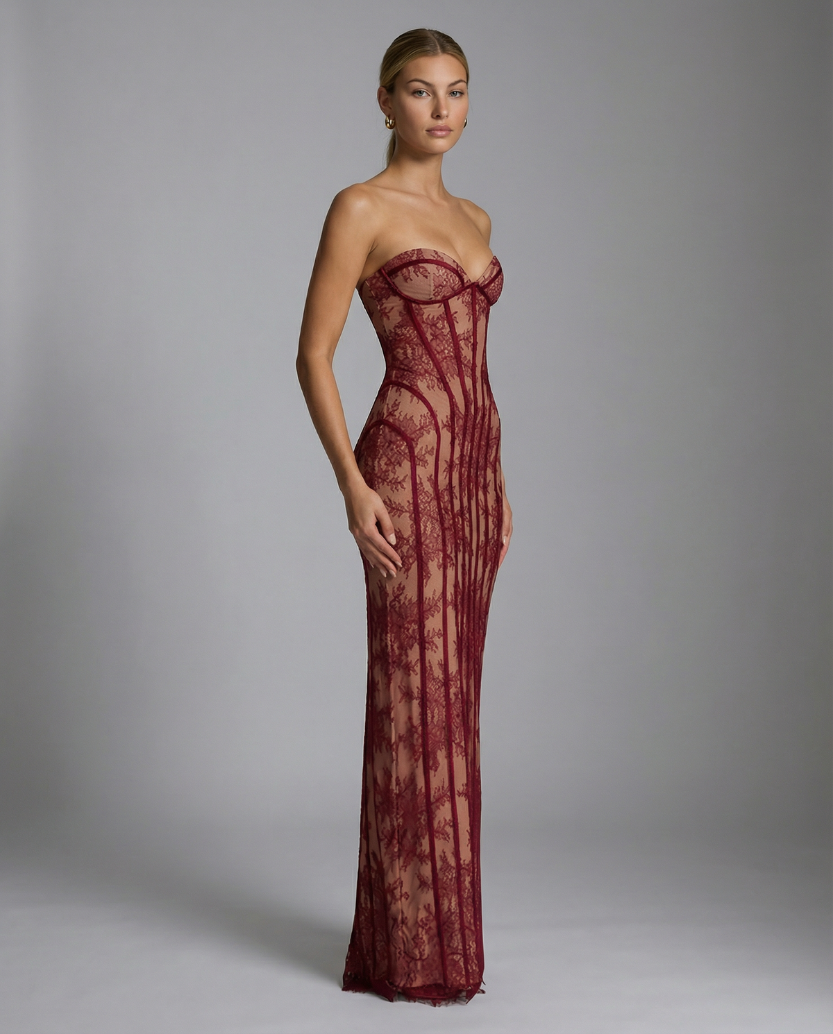 Side view of the burgundy strapless lace corset gown against a grey backdrop, showing the curved seams and floor-length skirt. 4