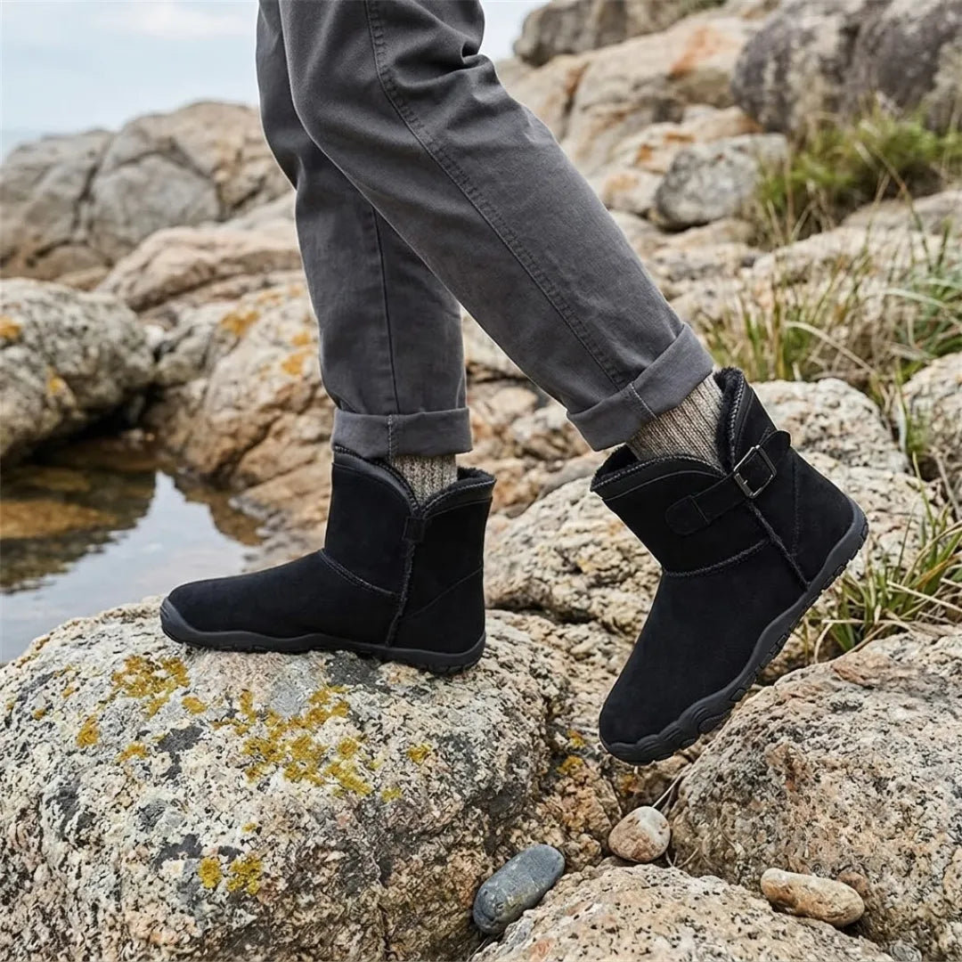 Black suede boots worn over patterned socks while stepping across lichen-covered rocks beside water 11