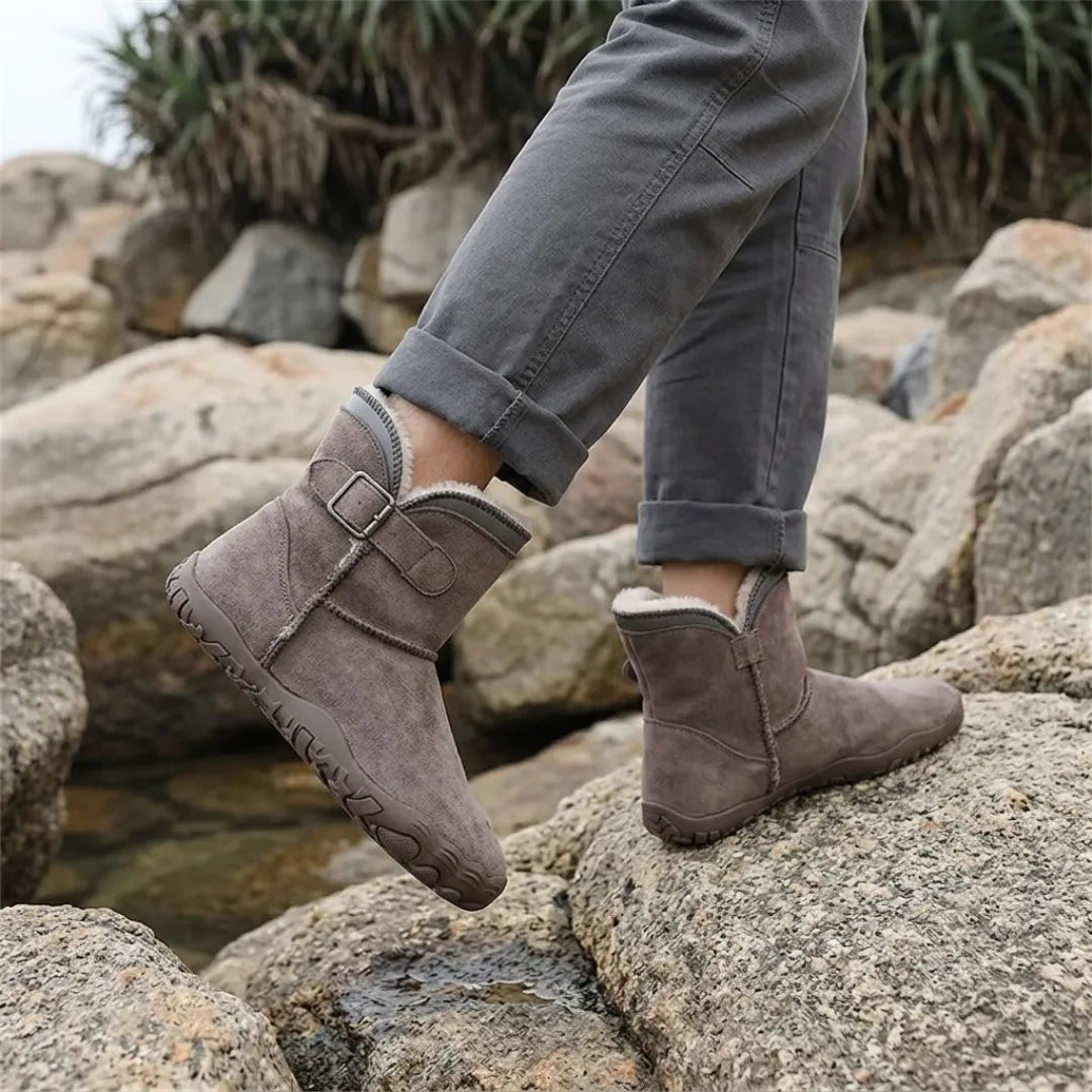 Model in rolled-up grey trousers wearing brown-grey fleece-lined boots while walking over large rocks by water 38