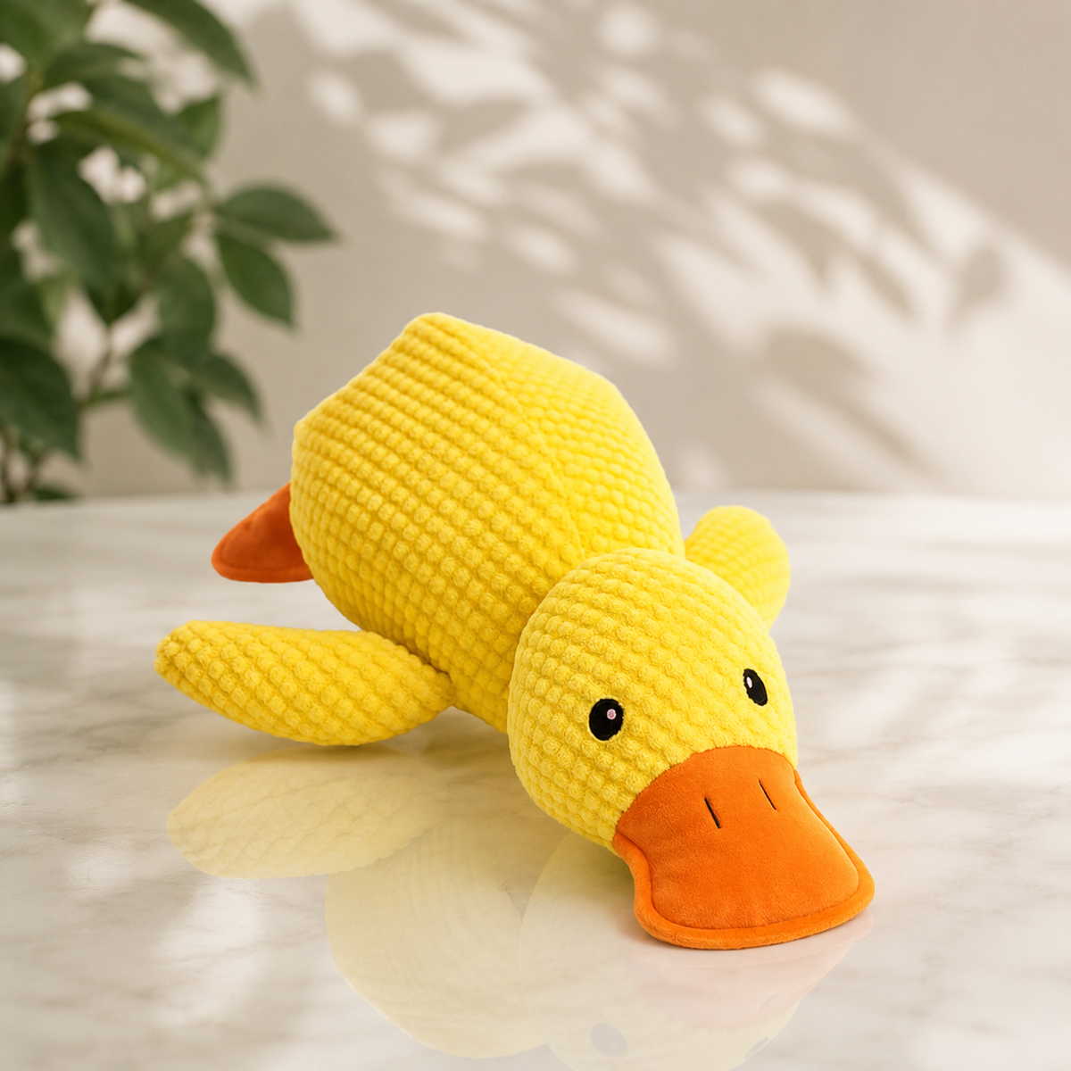 Yellow textured plush duck with orange bill lying on a marble surface beside a green plant 1