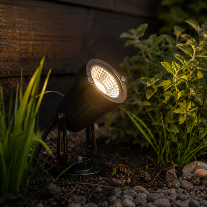 Angled view at dusk of the spotlight shining warm light, showing its adjustable bracket, cable and round base among plants and stones. 6