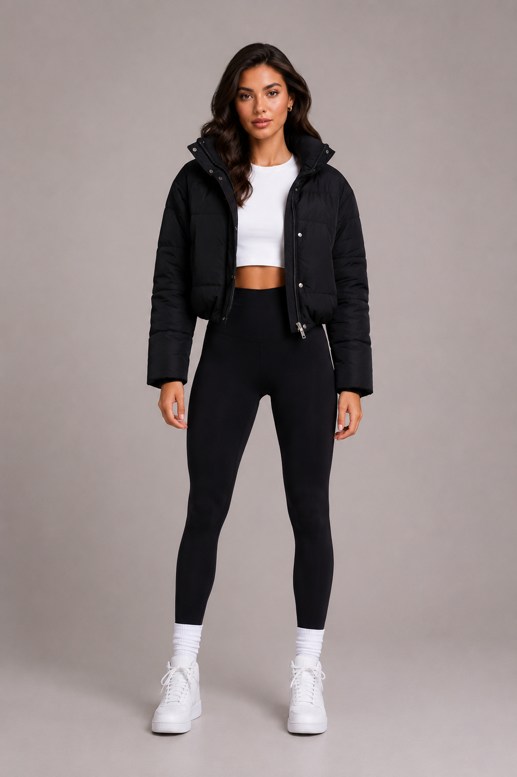Full-length front view of a model in the black cropped puffer jacket, white crop top, black leggings and white trainers, hood down. 2
