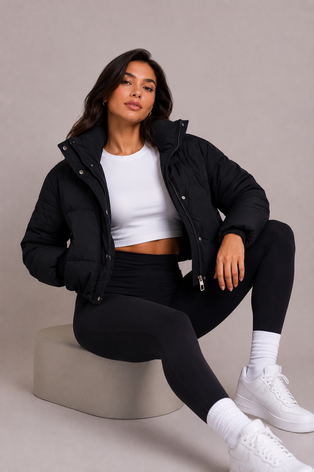 Model sitting on a stone block in the open black puffer jacket, white crop top, black leggings and white trainers. 6