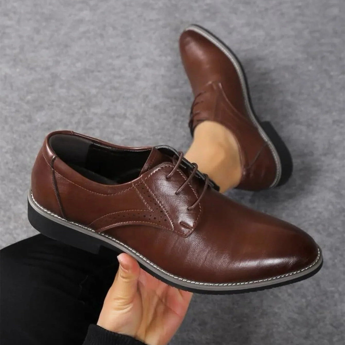 Dark brown polished lace-up shoe held in one hand above a grey carpet, with the second shoe resting beside it. 2