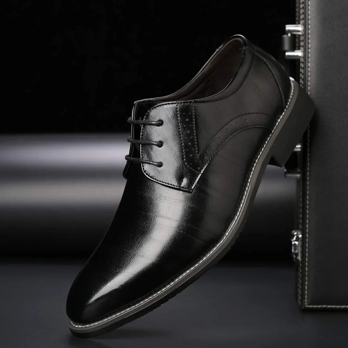 Single black glossy lace-up shoe with brogue perforations, tilted against a dark leather case on a dark studio background. 3