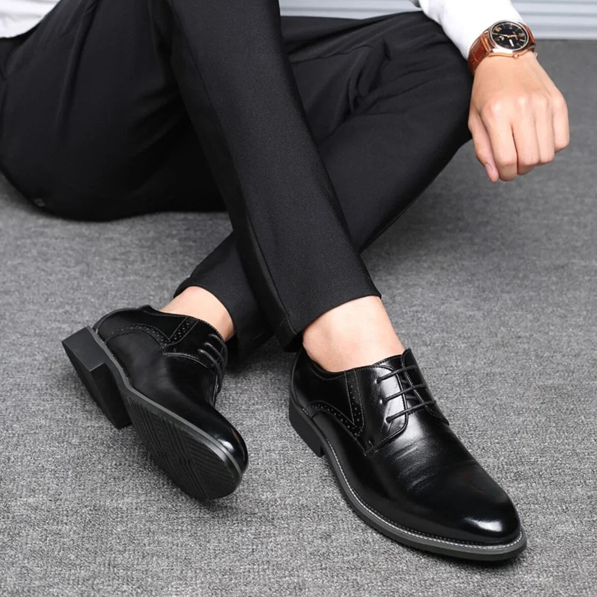 Man in black trousers sitting on grey carpet with legs crossed, wearing black polished lace-up shoes, one sole showing its tread. 4