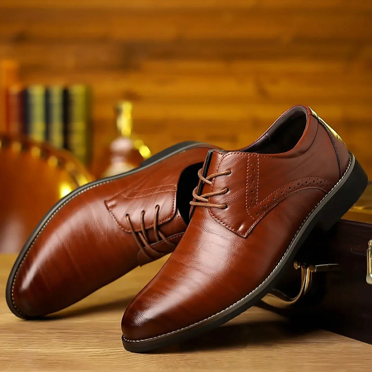 Pair of glossy tan-brown leather lace-up shoes with brogue detailing, resting at an angle on a wooden surface with a blurred bottle behind. 1
