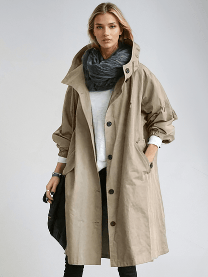 Model wears the long hooded coat in beige, buttoned loosely over a white top with a dark scarf and black trousers. 3
