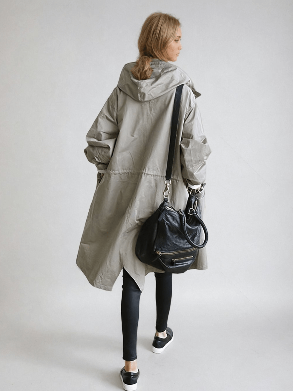 Back view of the long hooded coat in grey on a model, showing the hood, waist drawstring and a black shoulder bag. 4