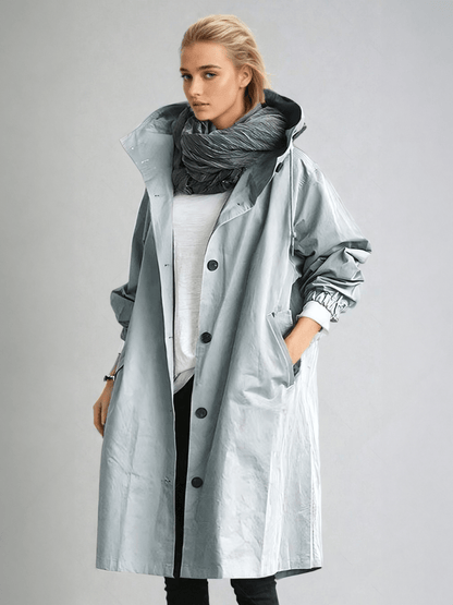 Model wears the long hooded coat in pale silvery blue-grey with a glossy finish, over white top and grey scarf, hand in pocket. 5