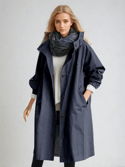 Model wears the long hooded coat in navy, with a high collar, front buttons and a dark scarf over a white jumper. 7