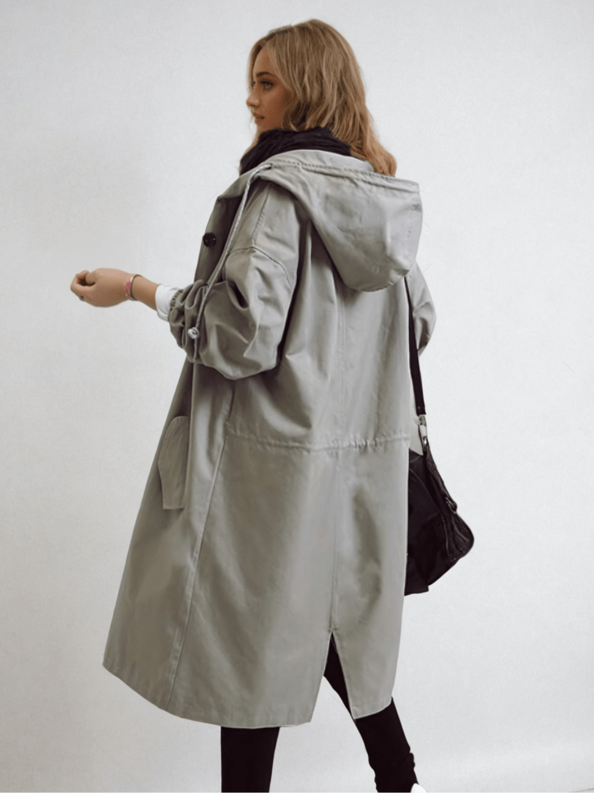 Rear three-quarter view of the long grey hooded coat showing the drawstring waist, back vent and a black bag over the shoulder. 8