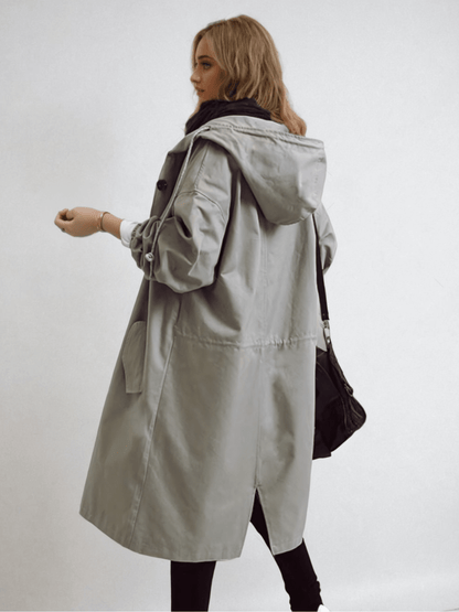 Rear three-quarter view of the long grey hooded coat showing the drawstring waist, back vent and a black bag over the shoulder. 8