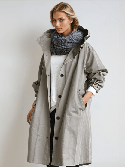 Model wears the long hooded coat in light grey, unbuttoned over a white top with a blue-grey scarf, one hand in pocket. 1