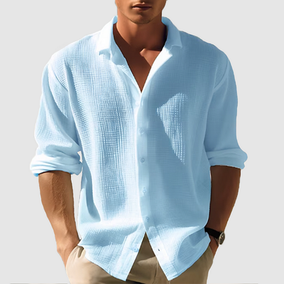 Model wearing the textured shirt in pale blue, sleeves rolled up, buttoned partway with a wide collar, hands in beige trouser pockets. 1