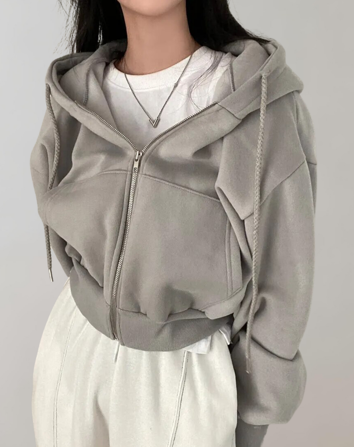 Grey cropped hoodie zipped up on a model, with braided drawstrings, front pockets and a ribbed hem, styled with white trousers. 4