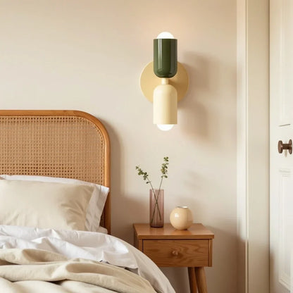 Dark green and cream wall lamp on a cream backplate beside a cane headboard, with a bedside table, glass vase and round ornament. 13