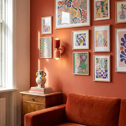 Deep red and peach wall lamp switched on among colourful framed prints in a terracotta room with a velvet sofa. 5
