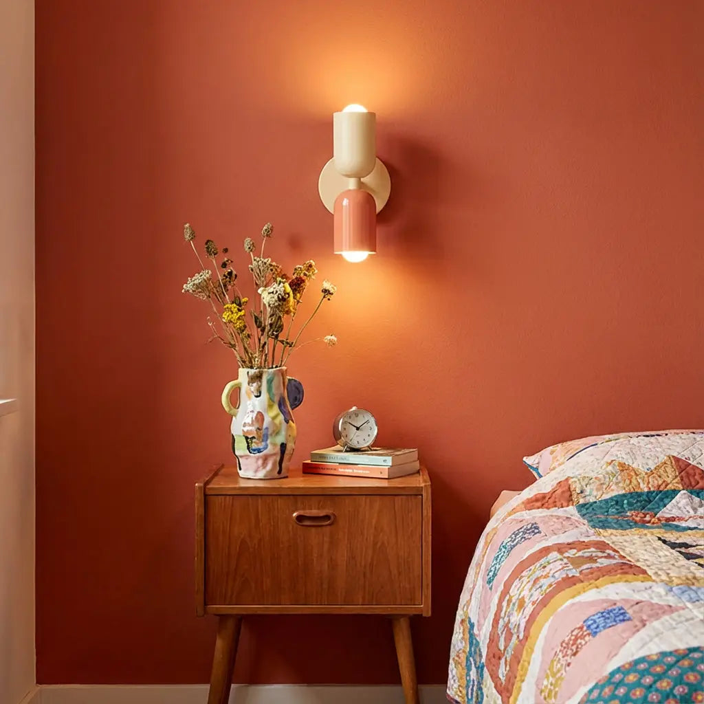 Cream and coral wall lamp glowing above a wooden bedside table, vase of dried flowers and a patchwork quilt, against a terracotta wall. 1