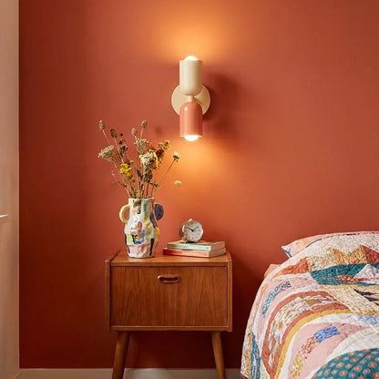Cream and coral wall lamp glowing above a wooden bedside table, vase of dried flowers and a patchwork quilt, against a terracotta wall. 1