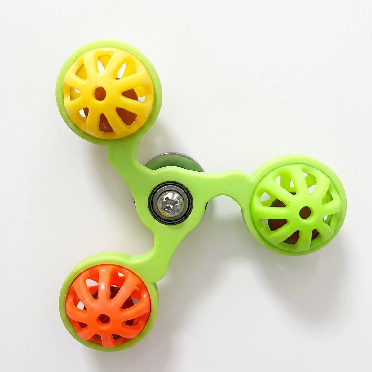 Three-armed spinner toy with a lime green frame, holding yellow, green and orange perforated plastic balls, shown flat against a plain white background. 1
