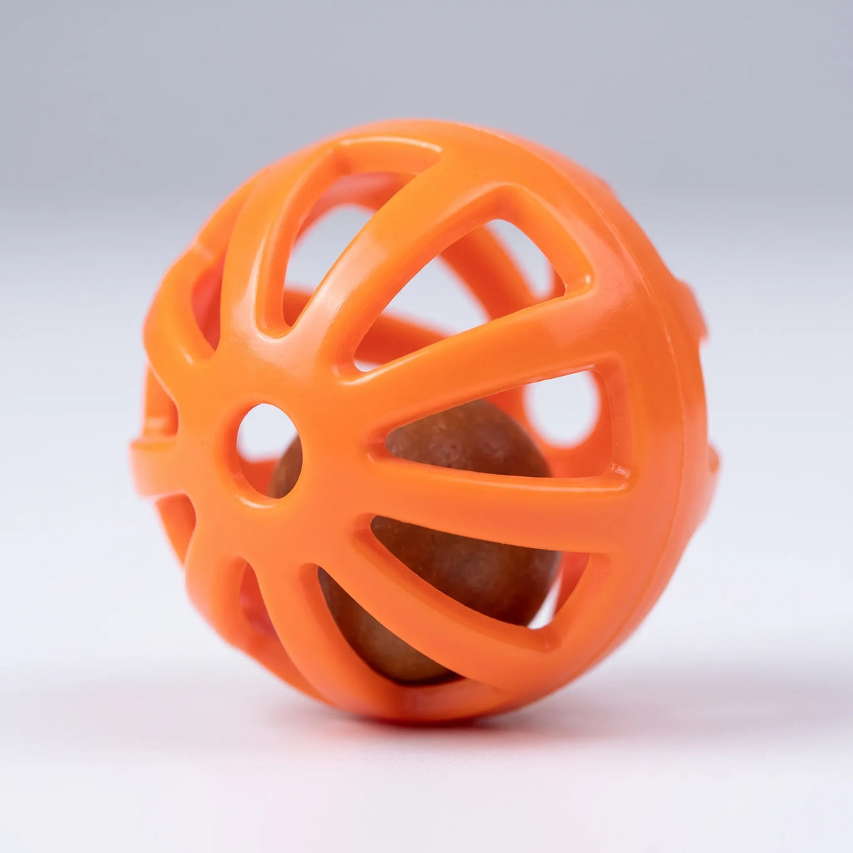 Close-up of a single orange perforated plastic ball with a small brown bead rattling inside, on a pale grey background. 4
