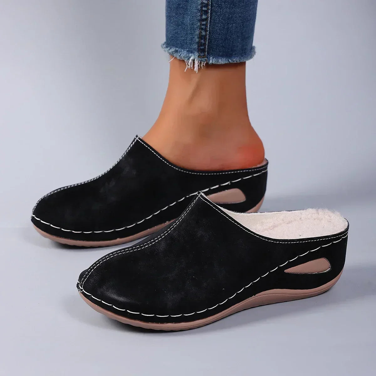 Black backless slippers worn by a model in frayed jeans, showing the fleece lining, stitched upper and cut-out wedge sole. 2