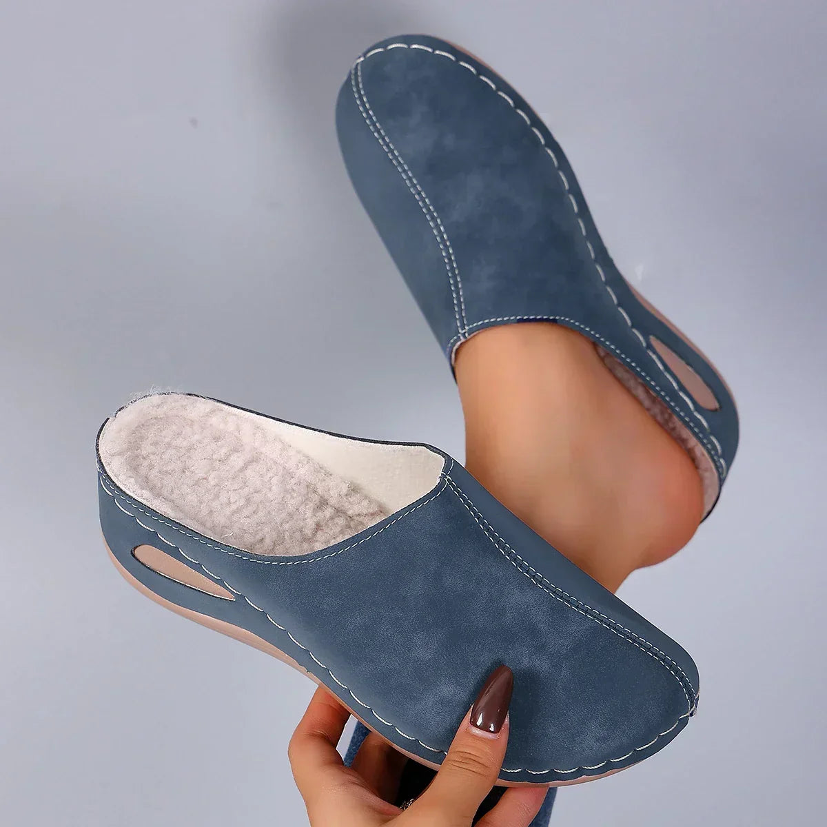 Pair of blue suede-look backless slippers held up to show the soft cream fleece lining and contrast stitching on the wedge sole. 1