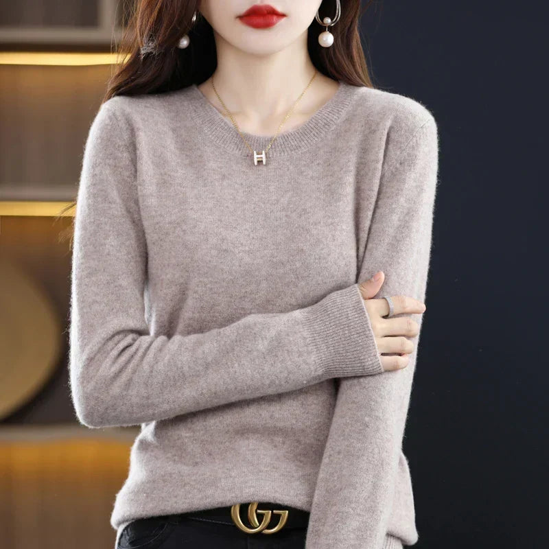 Light taupe crew-neck jumper worn with black trousers and a belt, one arm folded across the body 16