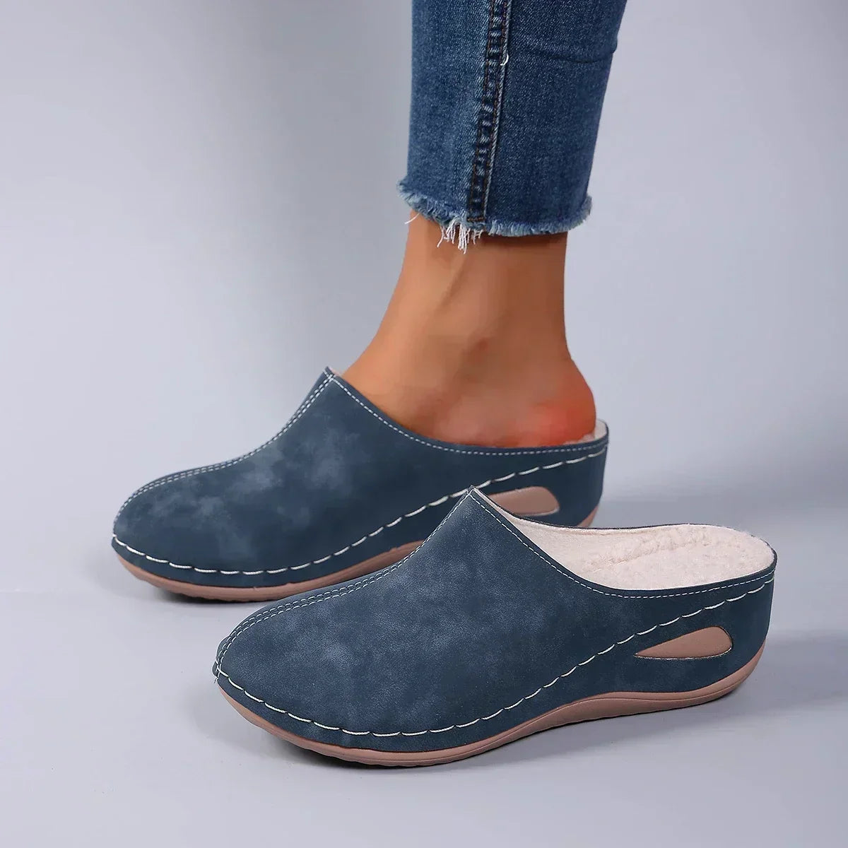 Blue backless slippers worn by a model in frayed jeans, showing the cream fleece lining and side cut-out in the sole. 4