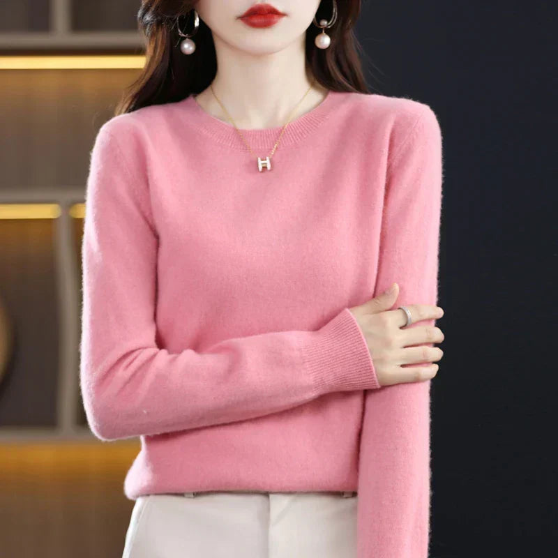 Pink crew-neck jumper with ribbed cuffs, worn with cream trousers and one arm folded across the body 10