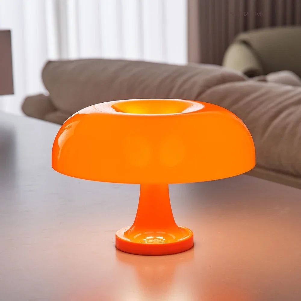 Close view of the orange lamp lit on a table, its glossy domed shade with a central dip in front of a sofa. 3