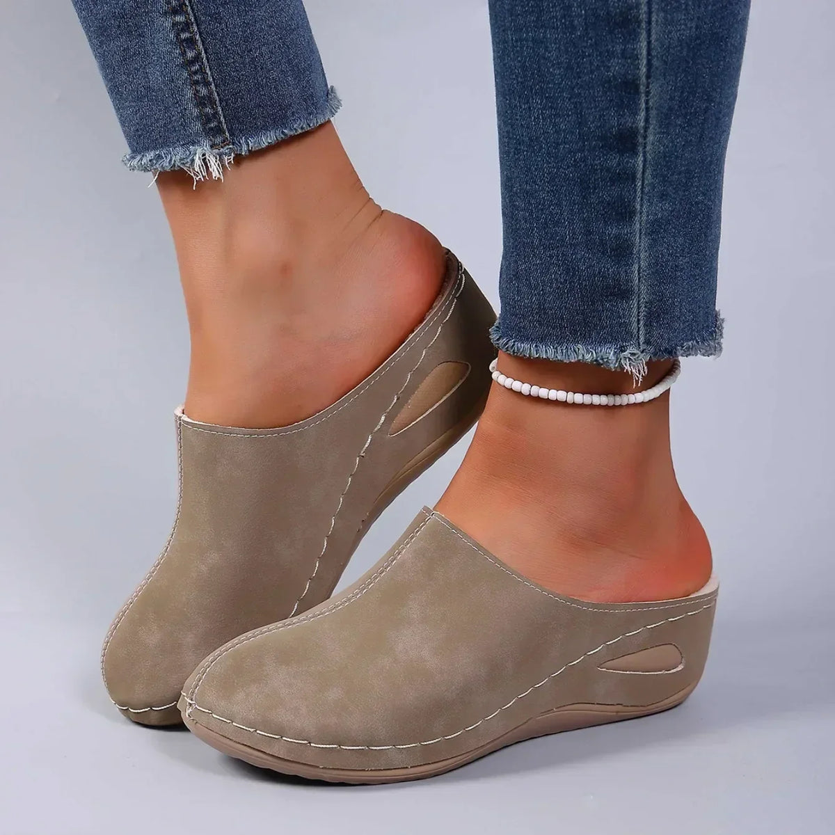 Taupe backless slippers on a model wearing frayed jeans and a beaded anklet, shown from the side with the cut-out wedge. 3