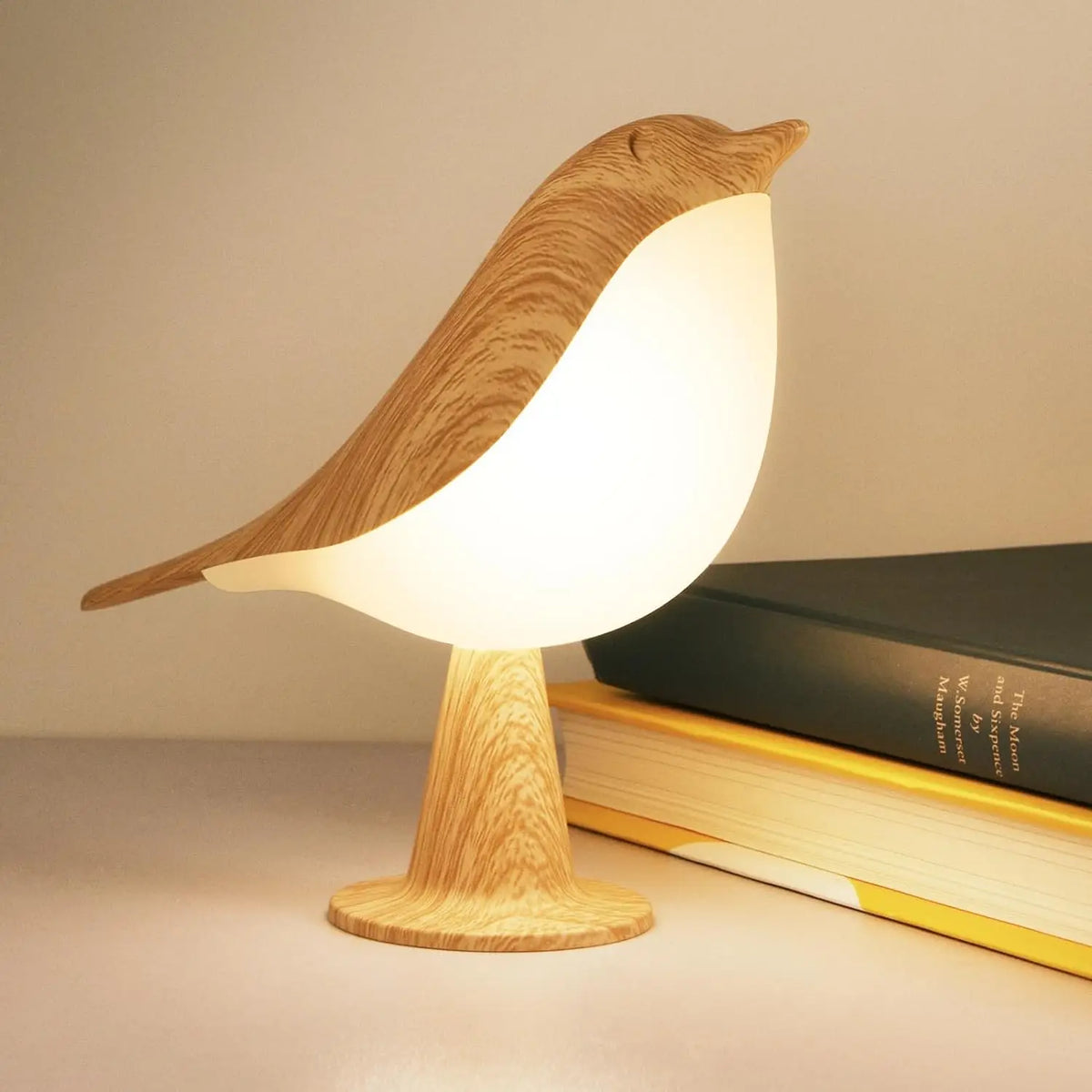 Bird-shaped lamp with a wood-effect head, tail and stem glowing warmly beside two stacked books 1
